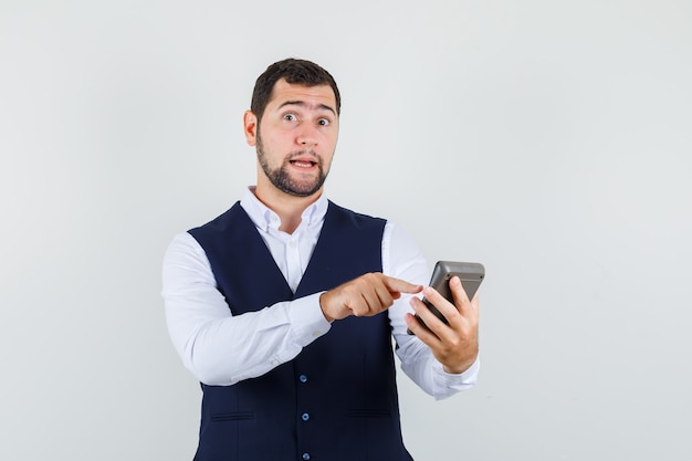 Jeune homme pointant sur la calculatrice en chemise, gilet et à hésitant
