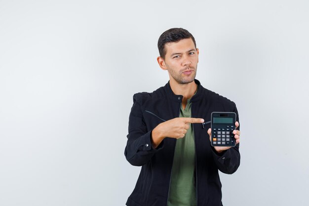 Jeune homme pointant sur la calculatrice en t-shirt, veste et semblant sensible, vue de face.