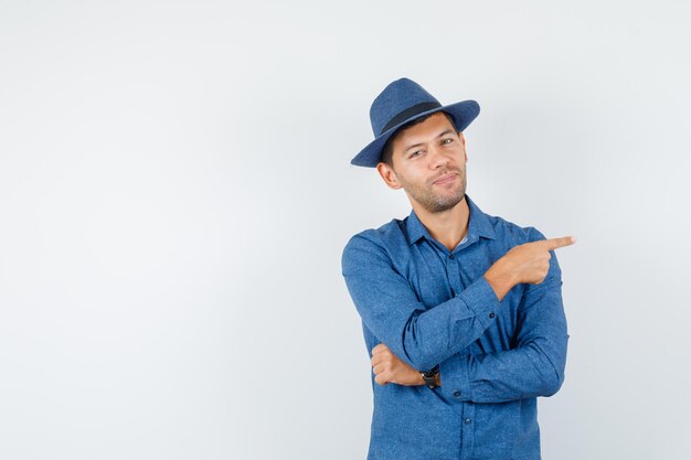 Jeune homme pointant sur le côté en chemise bleue, chapeau et à l'air confiant, vue de face.