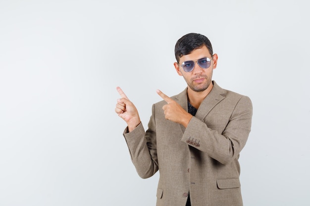 Jeune homme pointant de côté dans une veste marron grisâtre, des lunettes bleues et l'air assuré, vue de face.