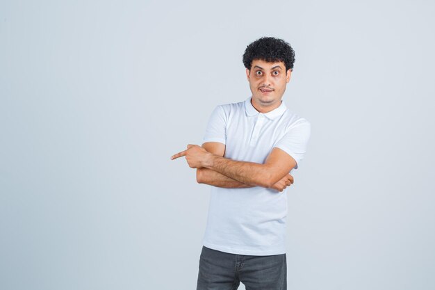 Jeune homme pointant de côté en t-shirt blanc, pantalon et l'air perplexe. vue de face.