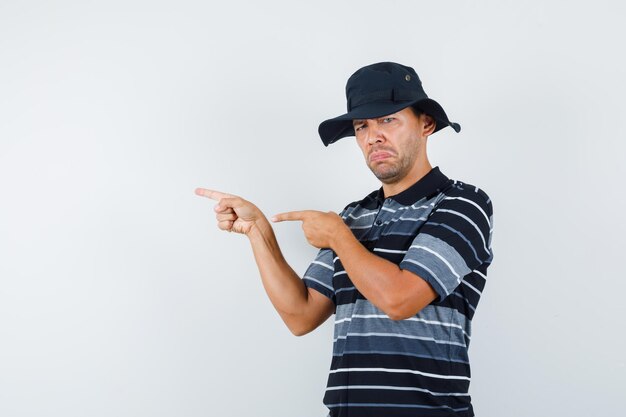 Jeune homme pointant sur le côté en t-shirt, chapeau et l'air triste, vue de face.
