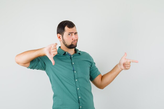 Jeune homme pointant de côté tout en montrant le pouce vers le bas en chemise verte et l'air insatisfait. vue de face.