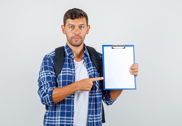 Jeune homme pointant le doigt sur le presse-papiers en chemise, sac à dos et à la grave, vue de face.