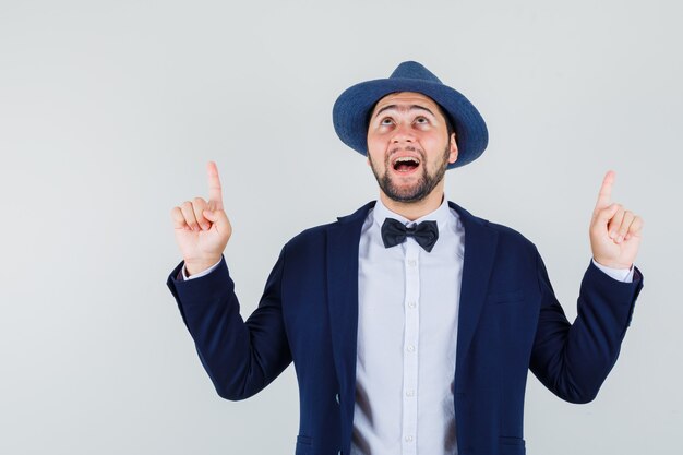 Jeune homme pointant les doigts vers le haut en costume, chapeau et à la recherche d'espoir, vue de face.