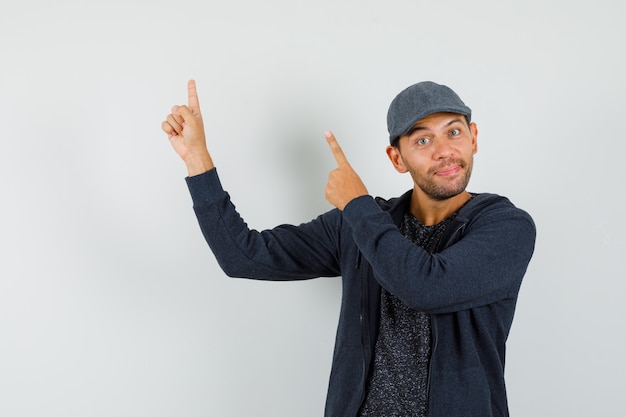 Jeune homme pointant les doigts vers le haut en t-shirt, veste, casquette et à la joyeuse.