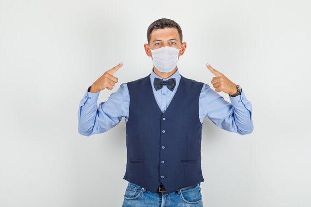 Jeune homme pointant du doigt son masque en costume, jeans et regardant prudemment.