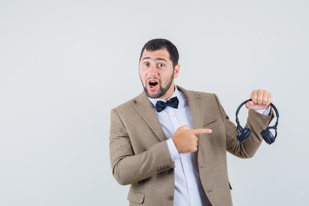 Jeune homme pointant sur les écouteurs en costume et à la vue positive, de face.