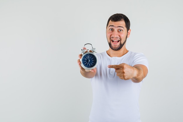 Jeune homme pointant sur l'horloge en t-shirt blanc et regardant jolly, vue de face. espace pour le texte