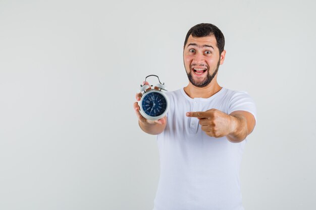 Jeune homme pointant sur l'horloge en t-shirt blanc et regardant jolly, vue de face. espace pour le texte