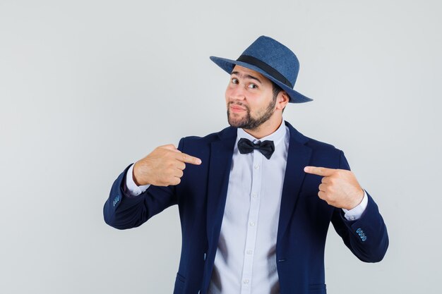 Jeune homme pointant sur lui-même en costume, chapeau et regardant beau, vue de face.