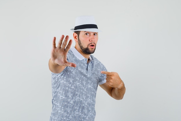 Jeune homme pointant sur lui-même tout en montrant le geste d'arrêt en t-shirt rayé, chapeau et à la troublé. vue de face.