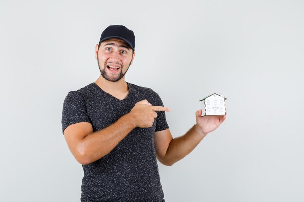 Jeune homme pointant sur le modèle de maison en t-shirt et casquette et à la bonne humeur