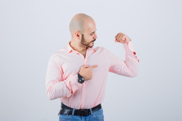 Jeune homme pointant les muscles du bras en chemise, jeans et l'air fier, vue de face.