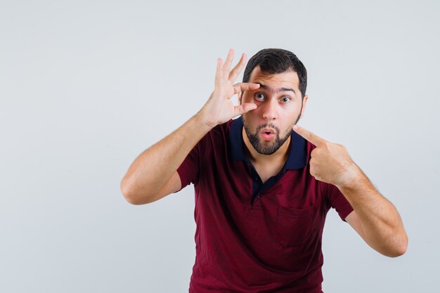 Jeune homme pointant sur sa paupière en t-shirt rouge et à la recherche concentrée. vue de face.