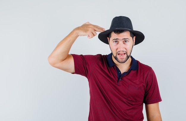 Jeune homme pointant sa tête avec signe de pistolet en t-shirt, chapeau et à la nervosité. vue de face.