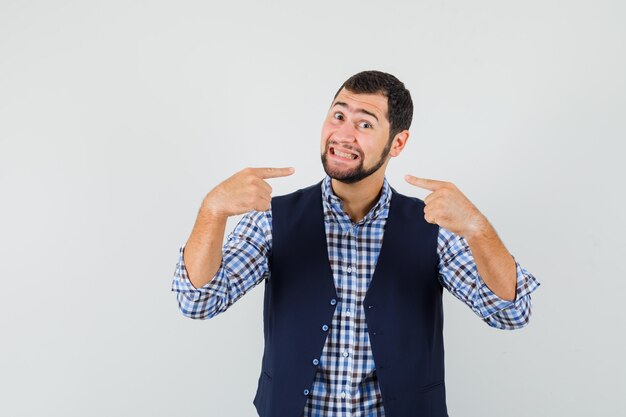 Jeune homme pointant sur ses dents en chemise, gilet et à la joyeuse vue de face.