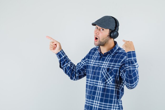 Jeune homme pointant sur ses écouteurs et de côté en chemise, casquette, vue de face.