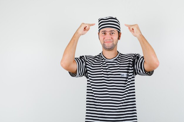 Jeune homme pointant sur son chapeau en t-shirt, chapeau et à la vue de face, confiant.