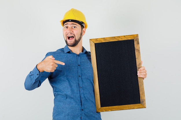 Jeune homme pointant sur tableau noir en chemise, casque et à l'optimiste, vue de face.