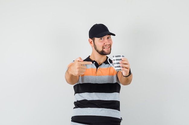 Jeune homme pointant sur une tasse de boisson en t-shirt, casquette et à la joyeuse. vue de face.