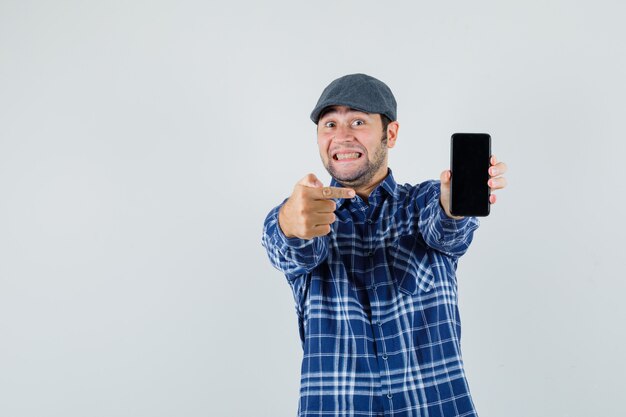 Jeune homme pointant sur téléphone mobile en chemise, casquette et à la vue de face, joyeuse.