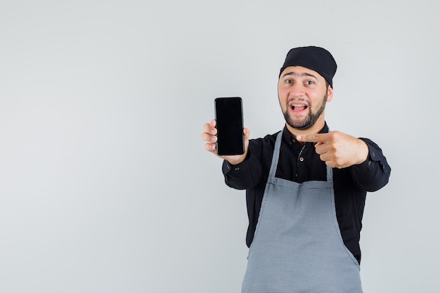 Jeune homme pointant sur téléphone mobile en chemise, tablier et à la vue de face, heureux.