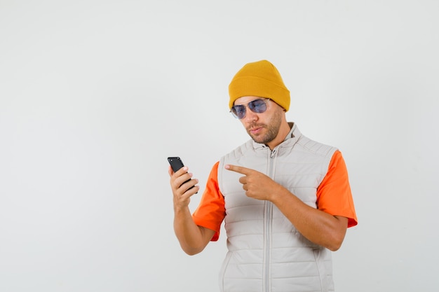 Jeune homme pointant sur téléphone mobile en t-shirt, veste, vue de face de chapeau.