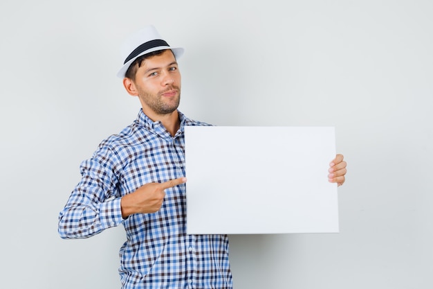 Jeune homme pointant sur une toile vide en chemise à carreaux, chapeau et à la recherche positive