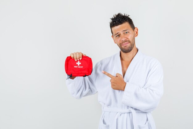 Jeune homme pointant sur une trousse de premiers soins en peignoir blanc, vue de face.