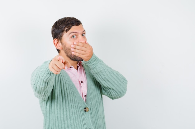 Jeune homme pointant vers l'avant, la main sur la bouche en chemise, cardigan et l'air surpris. vue de face.