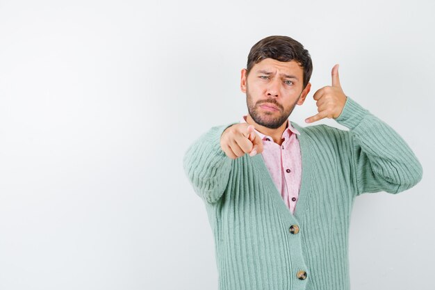 Jeune homme pointant vers l'avant tout en montrant le geste du téléphone en chemise, cardigan et ayant l'air confiant. vue de face.