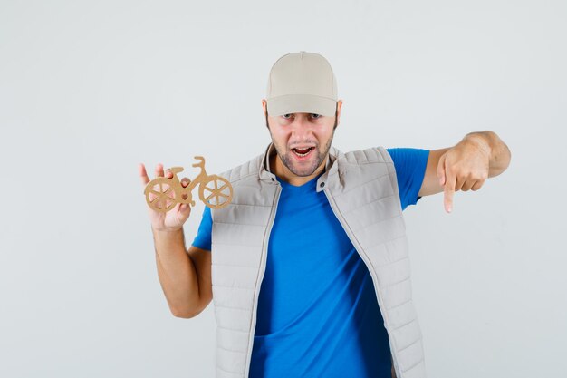 Jeune homme pointant vers le bas, tenant un vélo en bois en t-shirt, veste, casquette et à la confiance. vue de face.