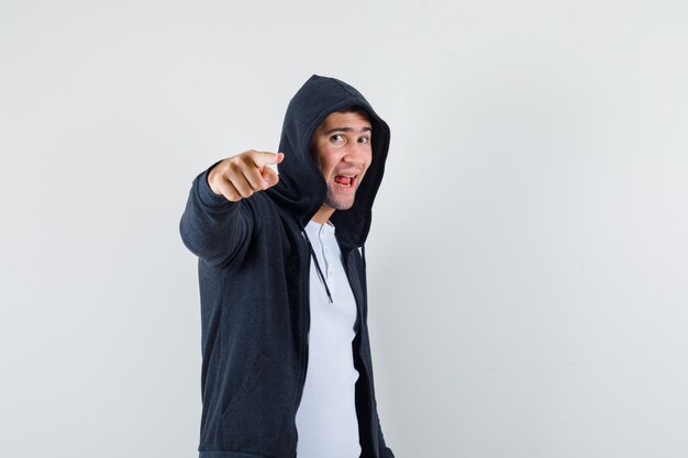 Jeune homme pointant vers la caméra en t-shirt, veste et regardant heureux, vue de face.