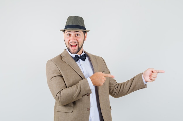 Jeune homme pointant vers le côté en costume, chapeau et à la joyeuse. vue de face.