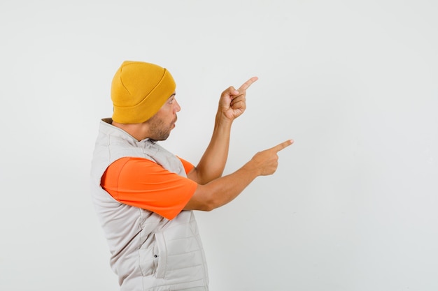 Jeune homme pointant vers le côté avec les doigts en t-shirt, veste, chapeau et à la surprise.