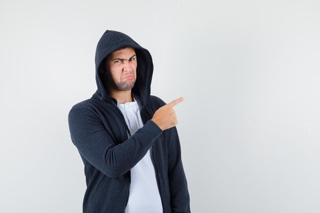 Jeune homme pointant vers le côté droit en t-shirt, veste et l'air mécontent. vue de face.