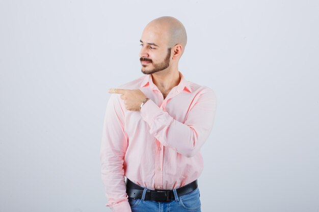 Jeune homme pointant vers le côté gauche en chemise, jeans et semblant confiant. vue de face.