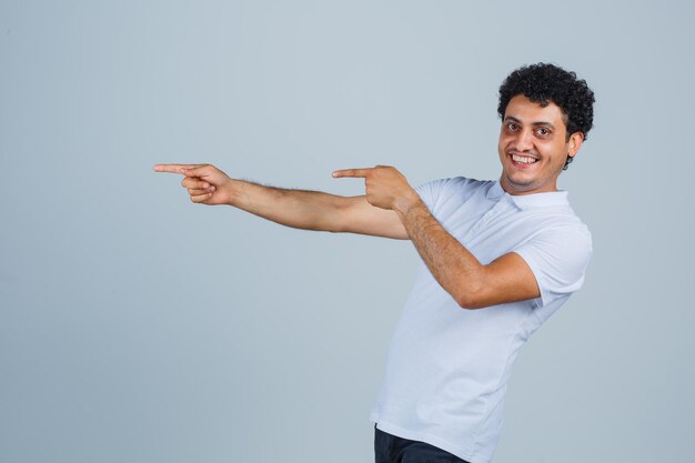 Jeune homme pointant vers le côté gauche en t-shirt blanc et regardant gai, vue de face.