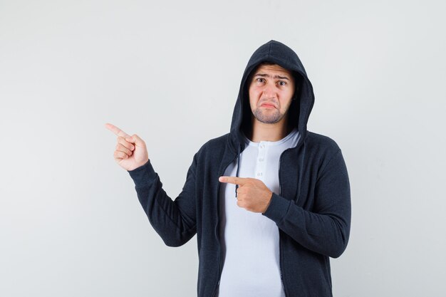 Jeune homme pointant vers le côté gauche en t-shirt, veste et à la triste. vue de face.