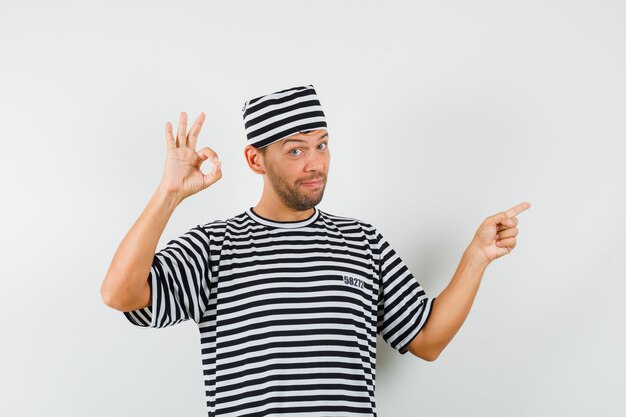 Jeune homme pointant vers le côté montrant le geste ok en chapeau t-shirt rayé et à la satisfaction