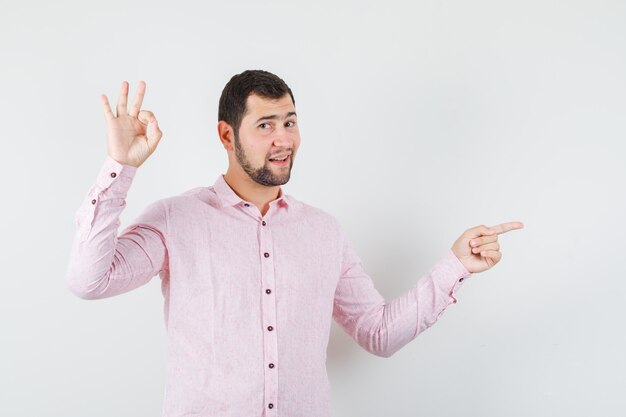Jeune homme pointant vers le côté avec signe ok en chemise rose et à la confiance