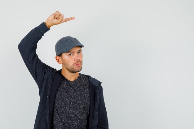 Jeune homme pointant vers le côté en t-shirt, veste, casquette et à la vue de face, hésitant.