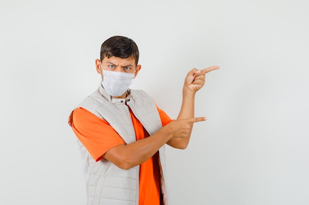 Jeune homme pointant vers le côté en t-shirt, veste, masque et à la recherche de sérieux.