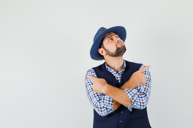 Jeune homme pointant vers l'extérieur en chemise, gilet, chapeau et à la vue réfléchie, de face.