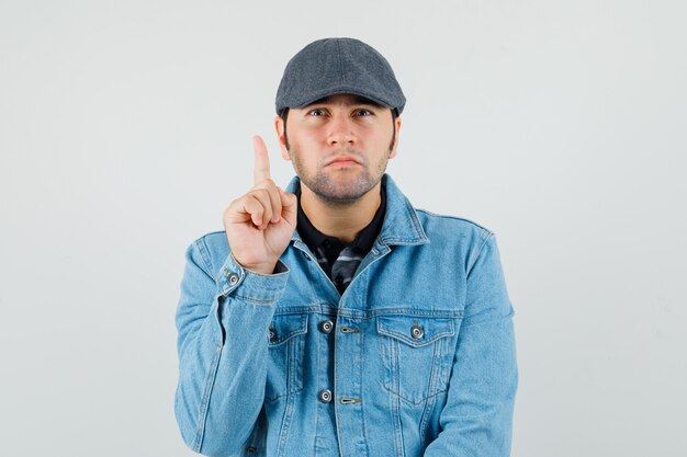 Jeune homme pointant vers le haut en casquette, t-shirt, veste et regardant curieux, vue de face.