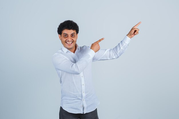 Jeune homme pointant vers le haut en chemise blanche, pantalon et l'air heureux. vue de face.