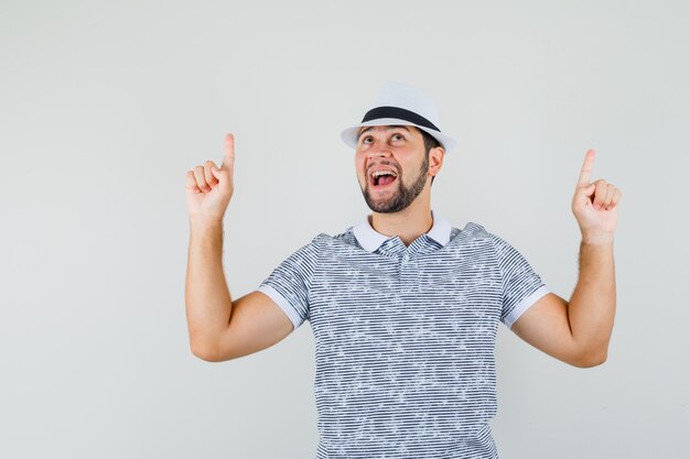 Jeune homme pointant vers le haut avec les doigts en t-shirt rayé, chapeau et à la vue de face, heureux.