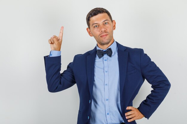 Jeune homme pointant vers le haut avec la main sur la taille en costume et à la joyeuse