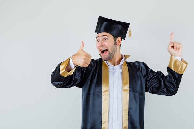 Jeune homme pointant vers le haut, montrant le pouce vers le haut en uniforme d'études supérieures et regardant heureux, vue de face.